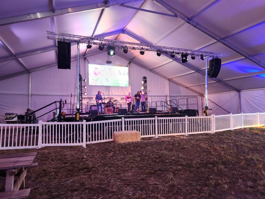 Band on a lit tent stage with truss lighting, flown speakers and a video screen